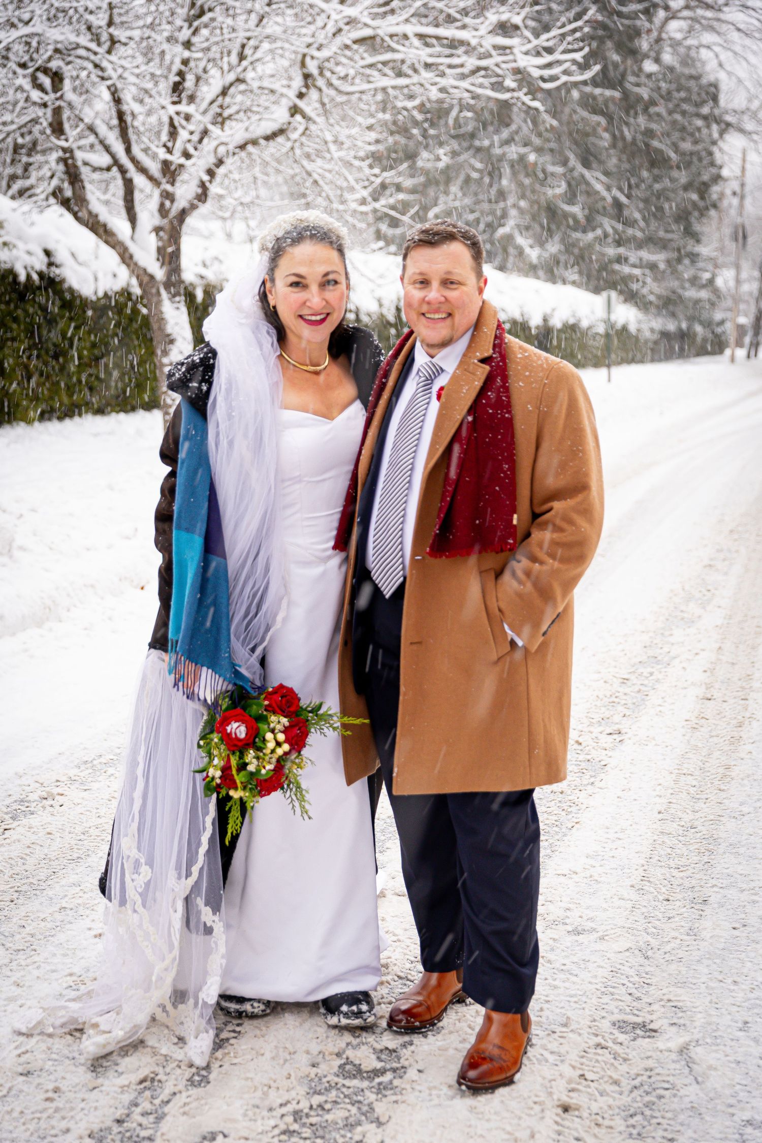 Bride and groom smiling together on a snowy tree-lined road, wearing winter coats over wedding attire.