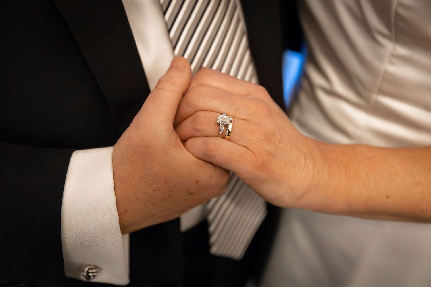 Close-up of bride and groom holding hands, showcasing a diamond engagement ring and wedding band.