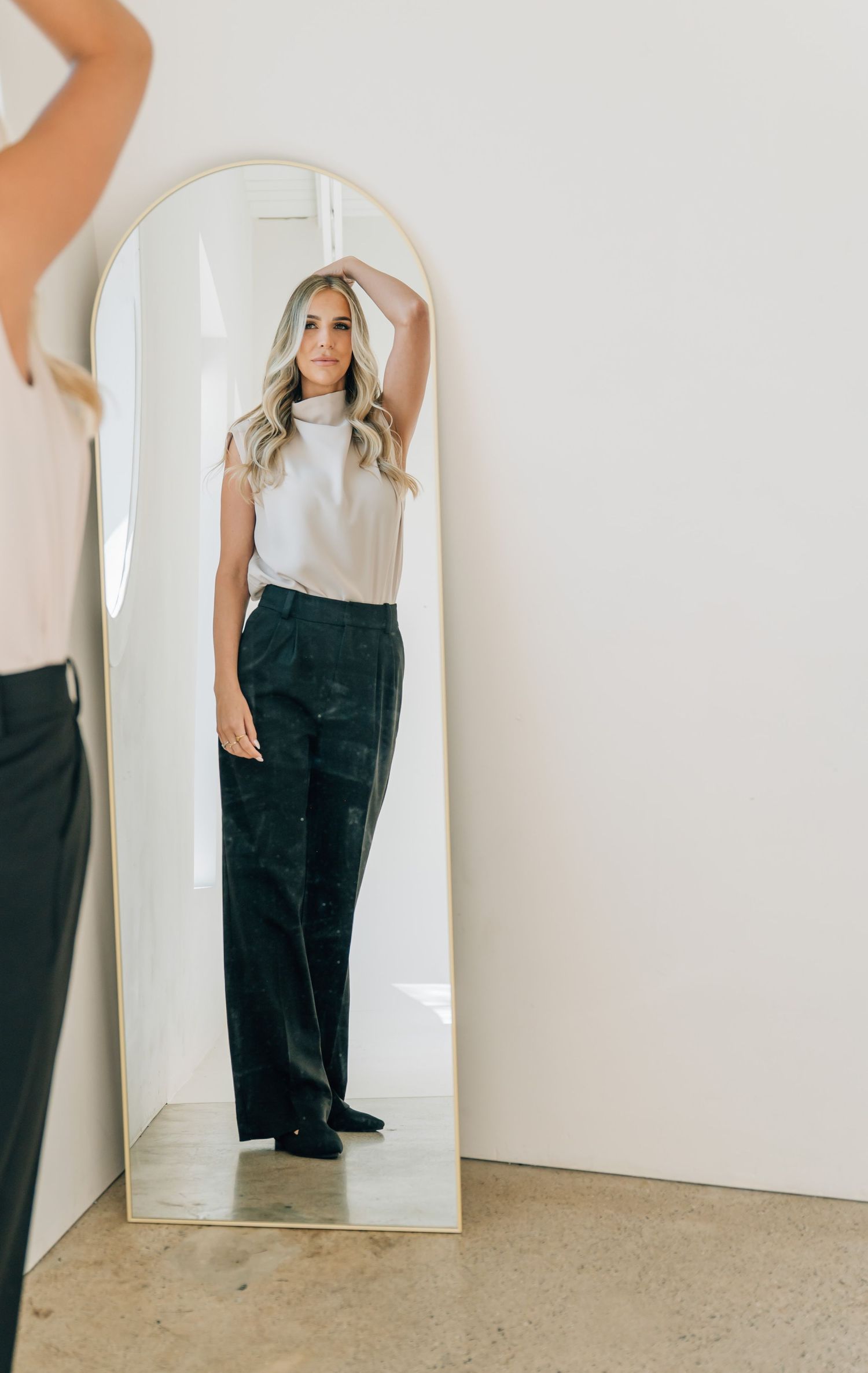 Mirror reflection showing a casual outfit with a white sleeveless top and dark pants against a white wall.