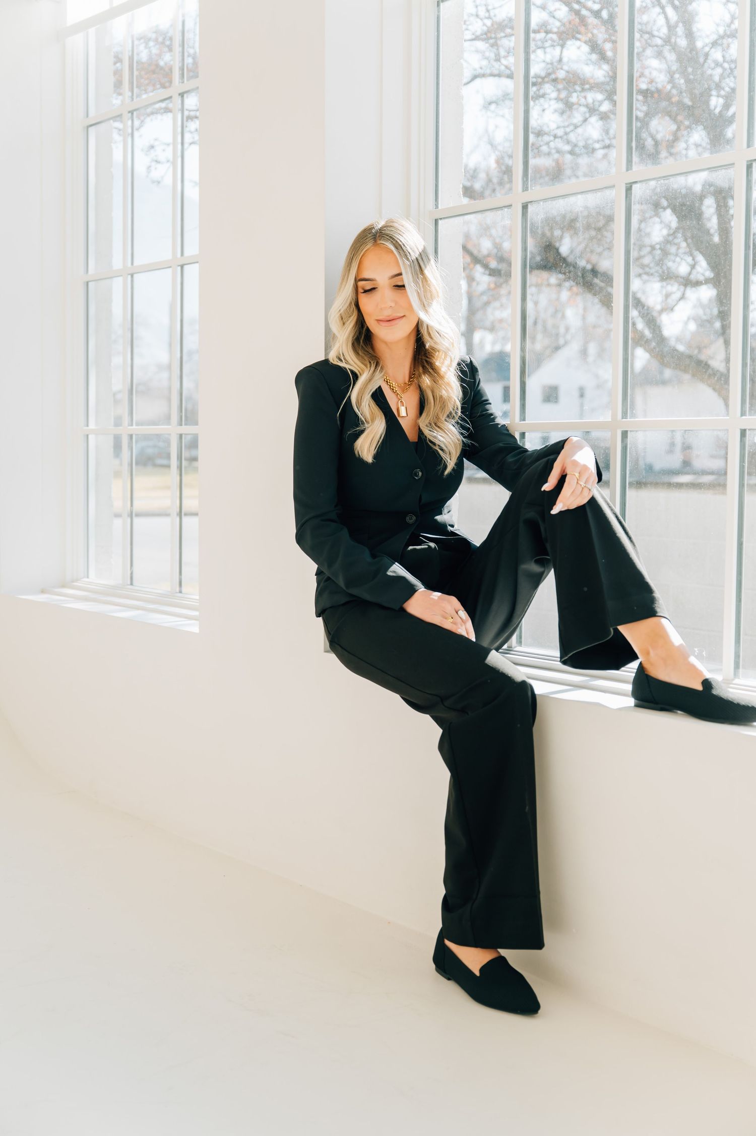 Fashion model in black blazer dress poses elegantly against bright white interior with large windows.