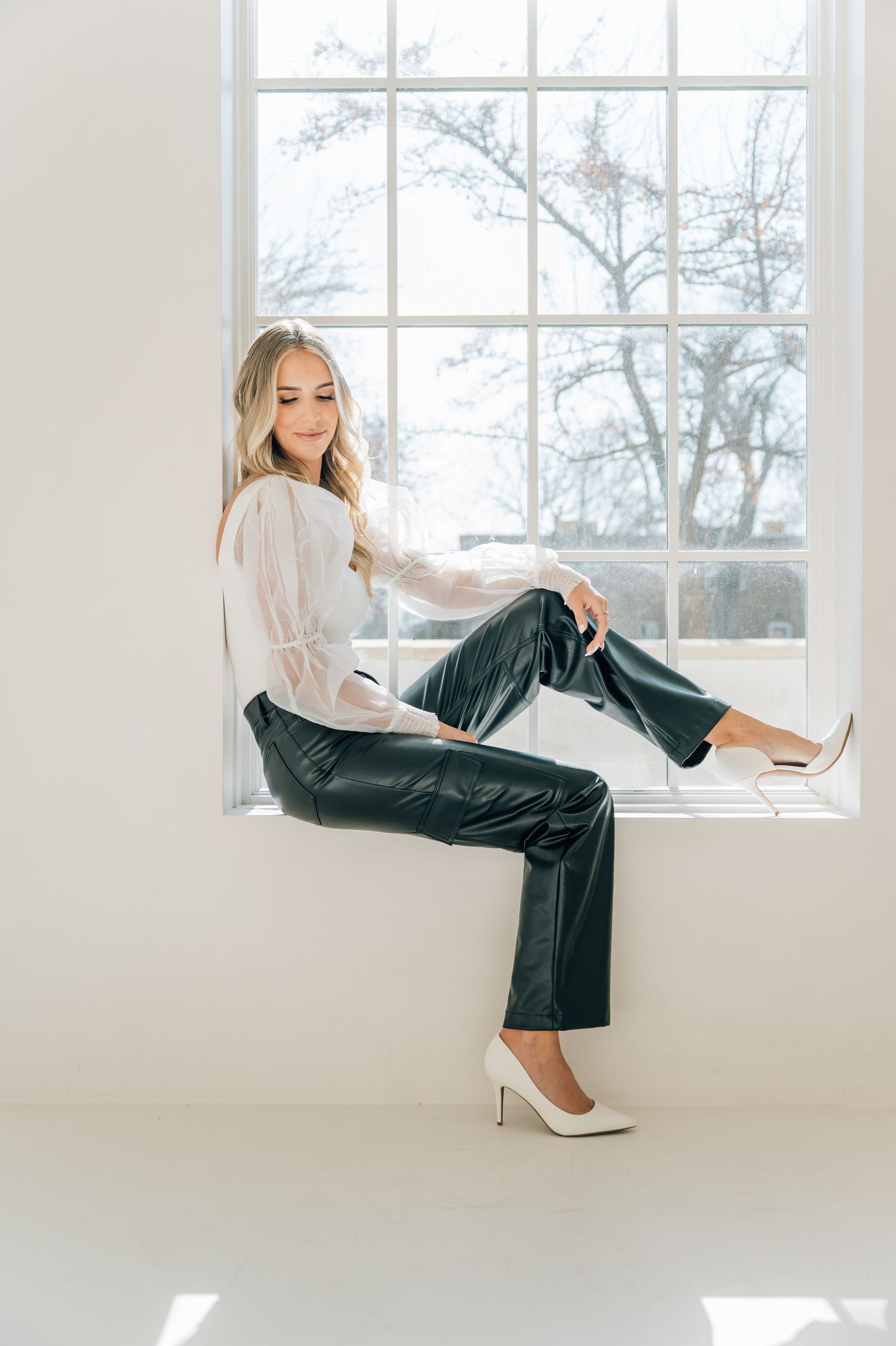 A woman in a white blouse and black leather pants poses gracefully near a bright window in a minimalist white room.
