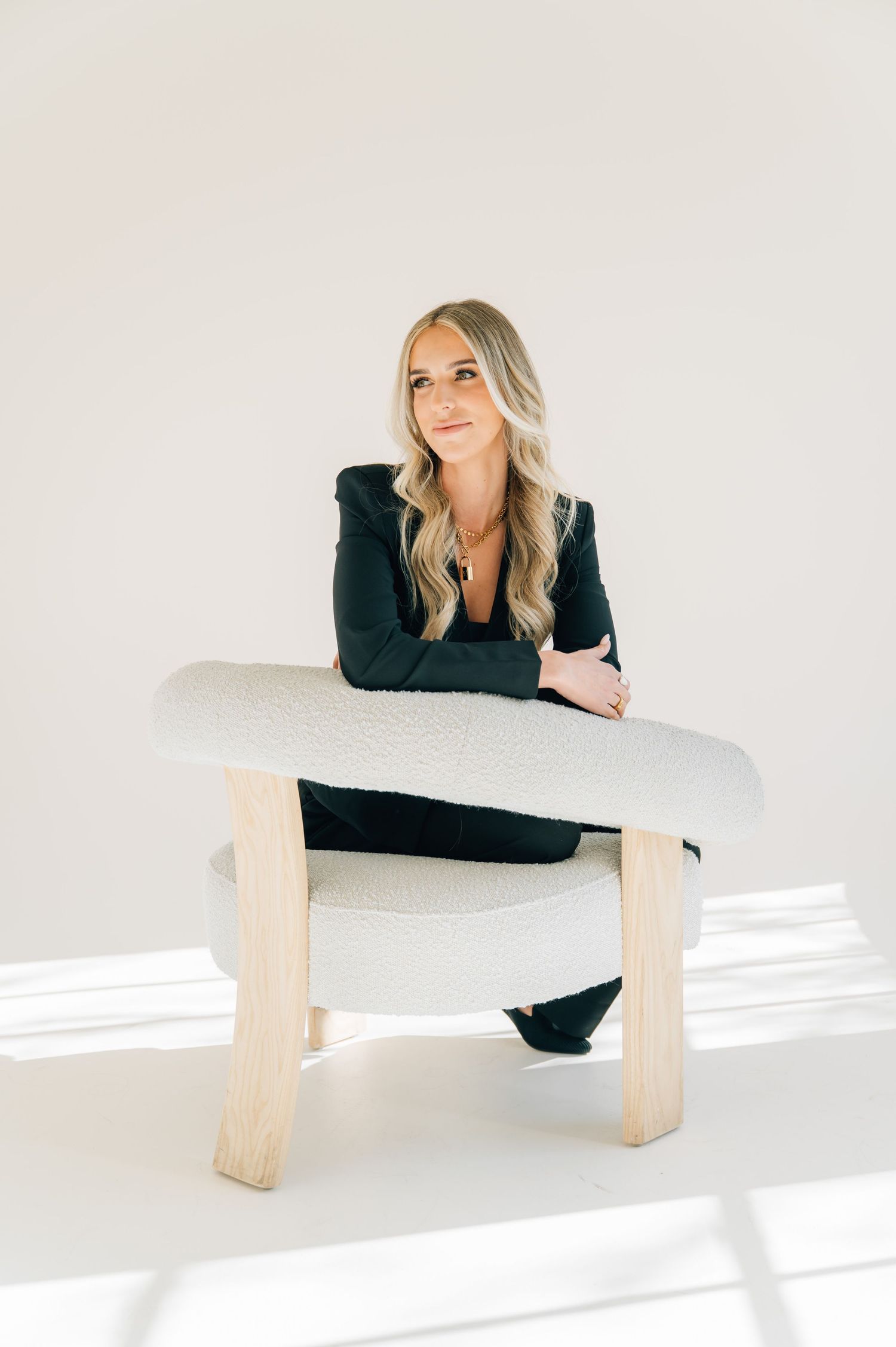 A person in a black outfit poses on a modern wooden bench in a bright minimalist white studio setting.