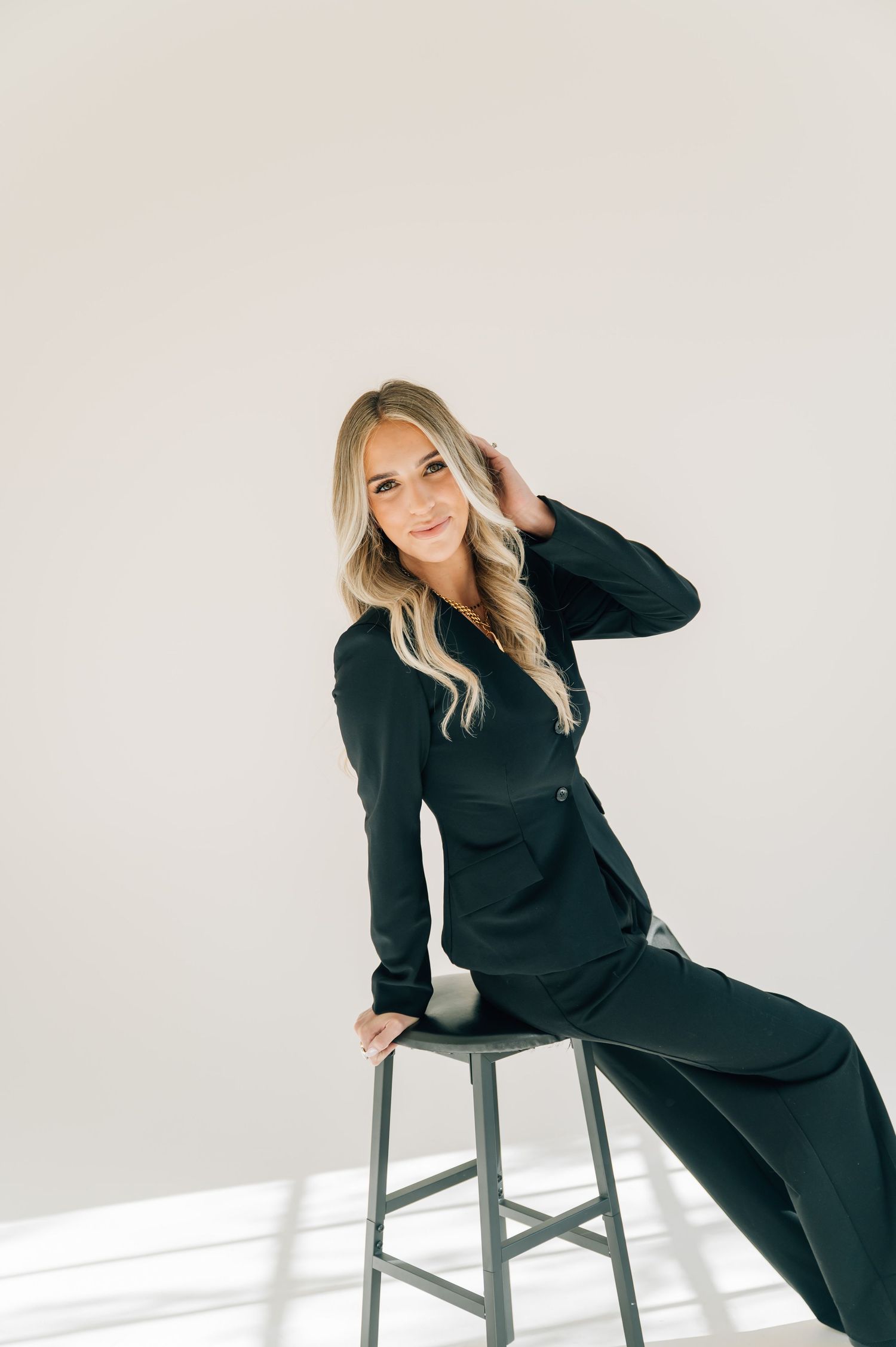Stylish woman in black outfit poses on grey stool in minimalist white studio setting.