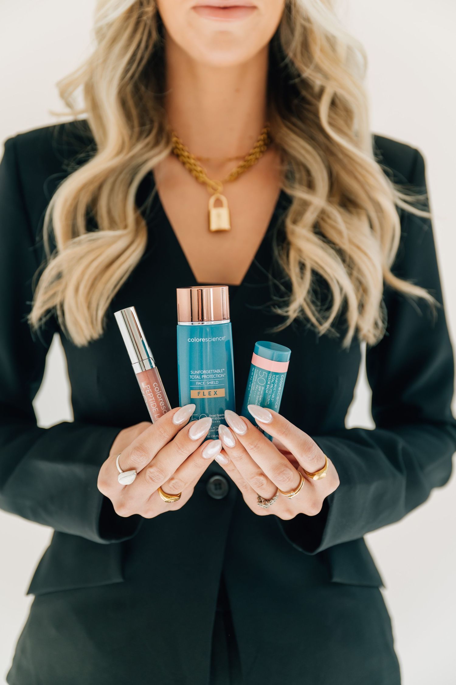 Woman in black blazer holding beauty products wearing gold chain necklace with padlock pendant.