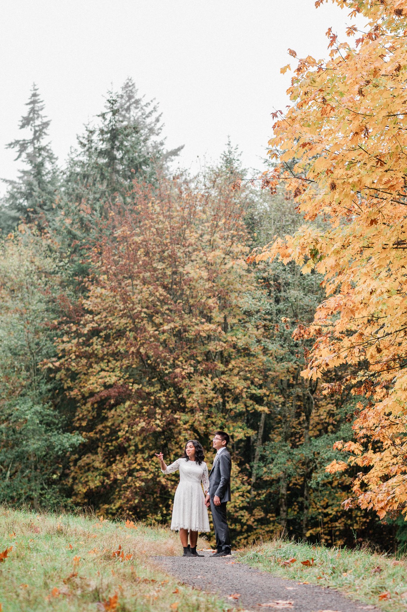 A bride and groom stands on a forest path surrounded by vibrant autumn foliage and evergreen trees on a misty fall day.