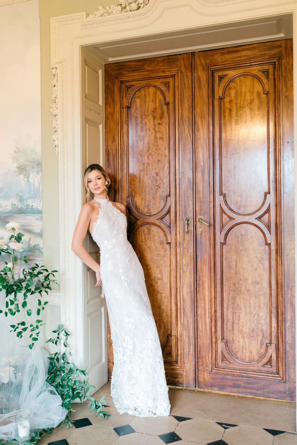 Elegant white lace wedding gown photographed against ornate wooden doors with decorative wall murals and greenery.