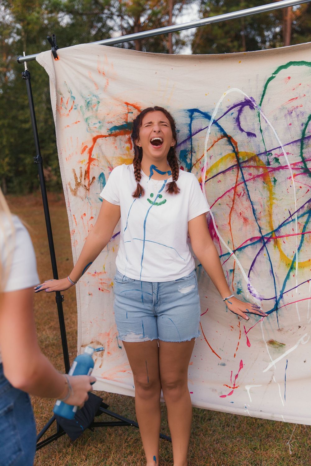 A person in white shirt and denim shorts stands in front of a paint-splattered white canvas outdoors.