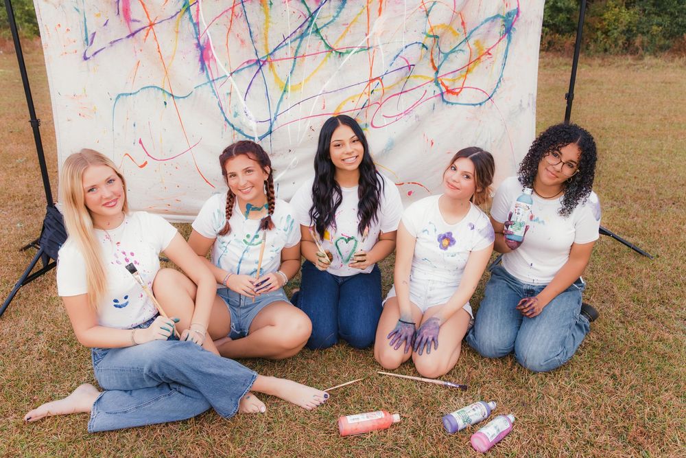 A group of friends in casual attire sitting together outdoors on grass while holding drinks and snacks.