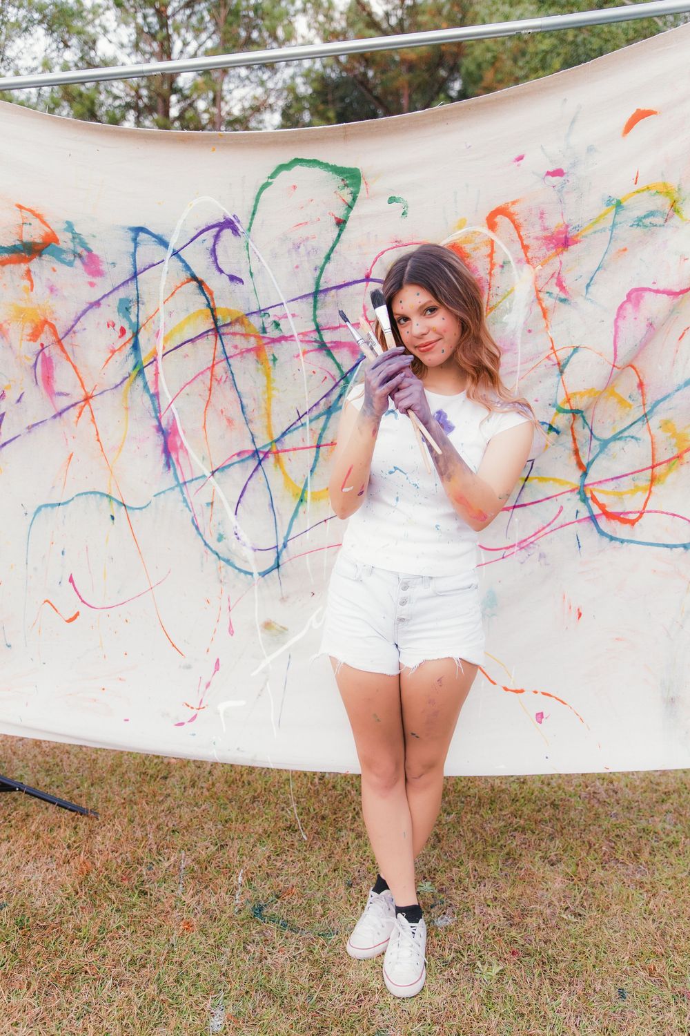 A woman in white attire poses creatively in front of a wall decorated with colorful paint splatter art.