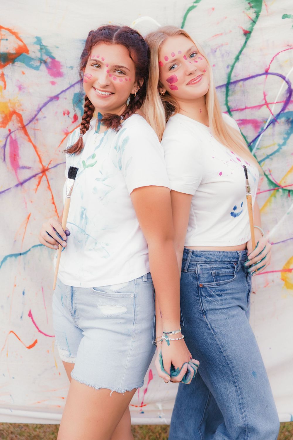 Two friends in white tops and denim pose playfully against a colorful paint splattered wall.