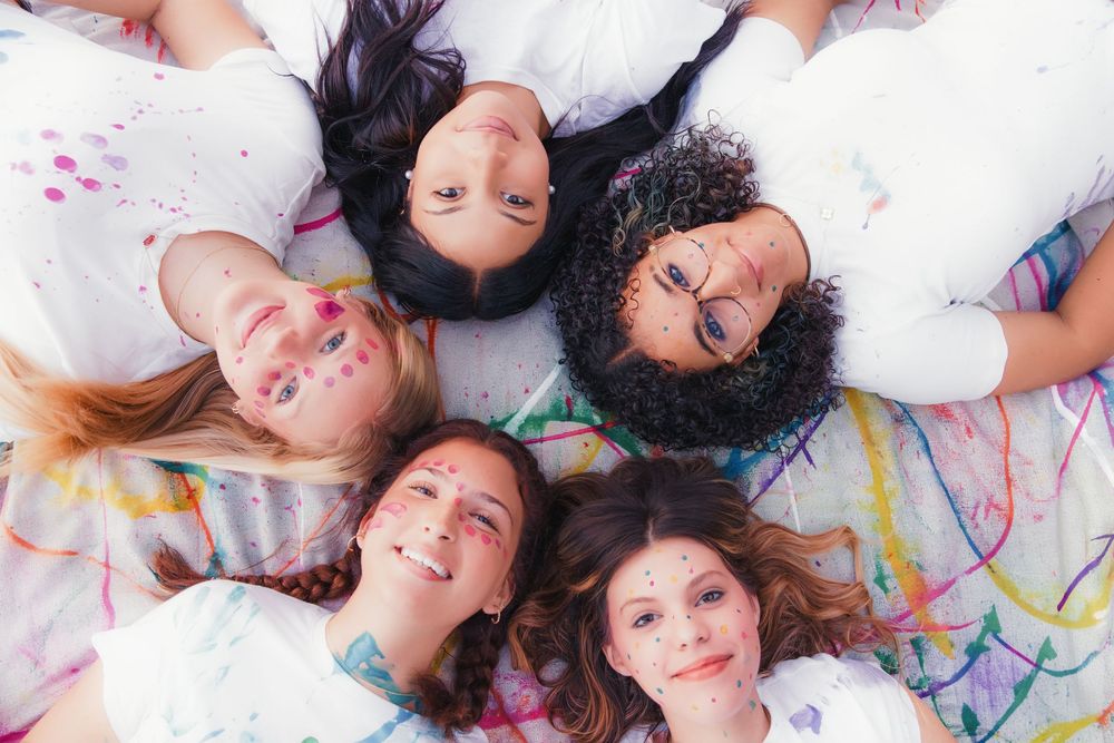 People lying on white surface with colorful paint marks on their faces smiling and laughing together.