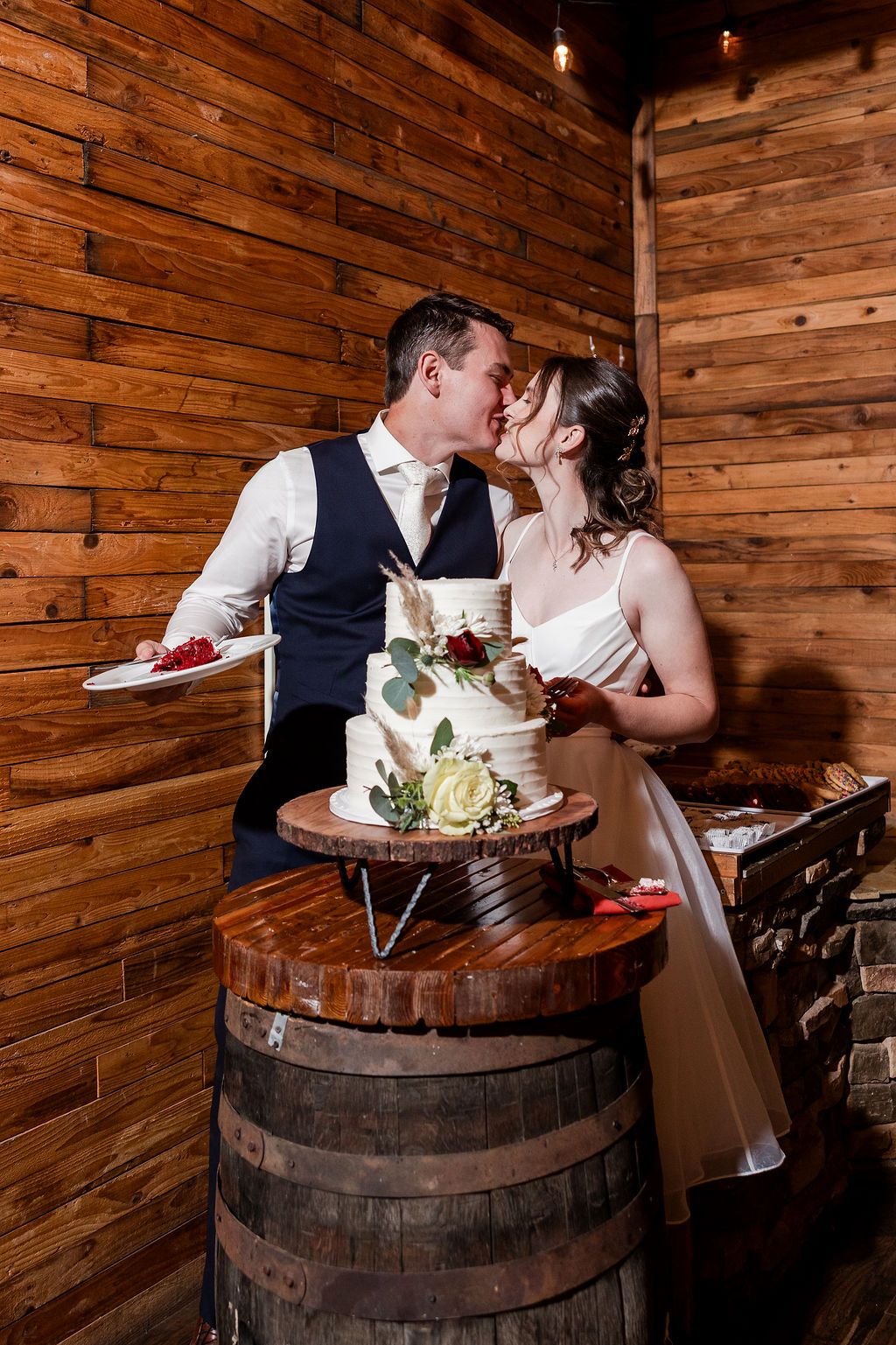Couple shares romantic moments while enjoying cake on rustic wooden wine barrel in barn-style setting.