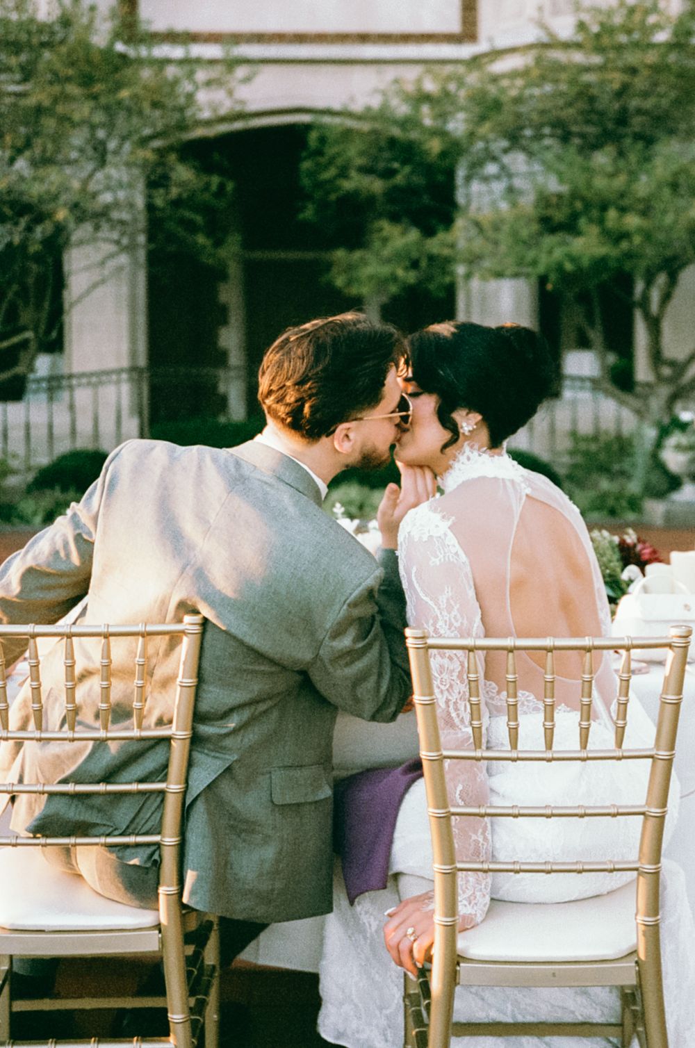 A couple sitting in white chiavari chairs share a romantic kiss at an outdoor wedding reception.