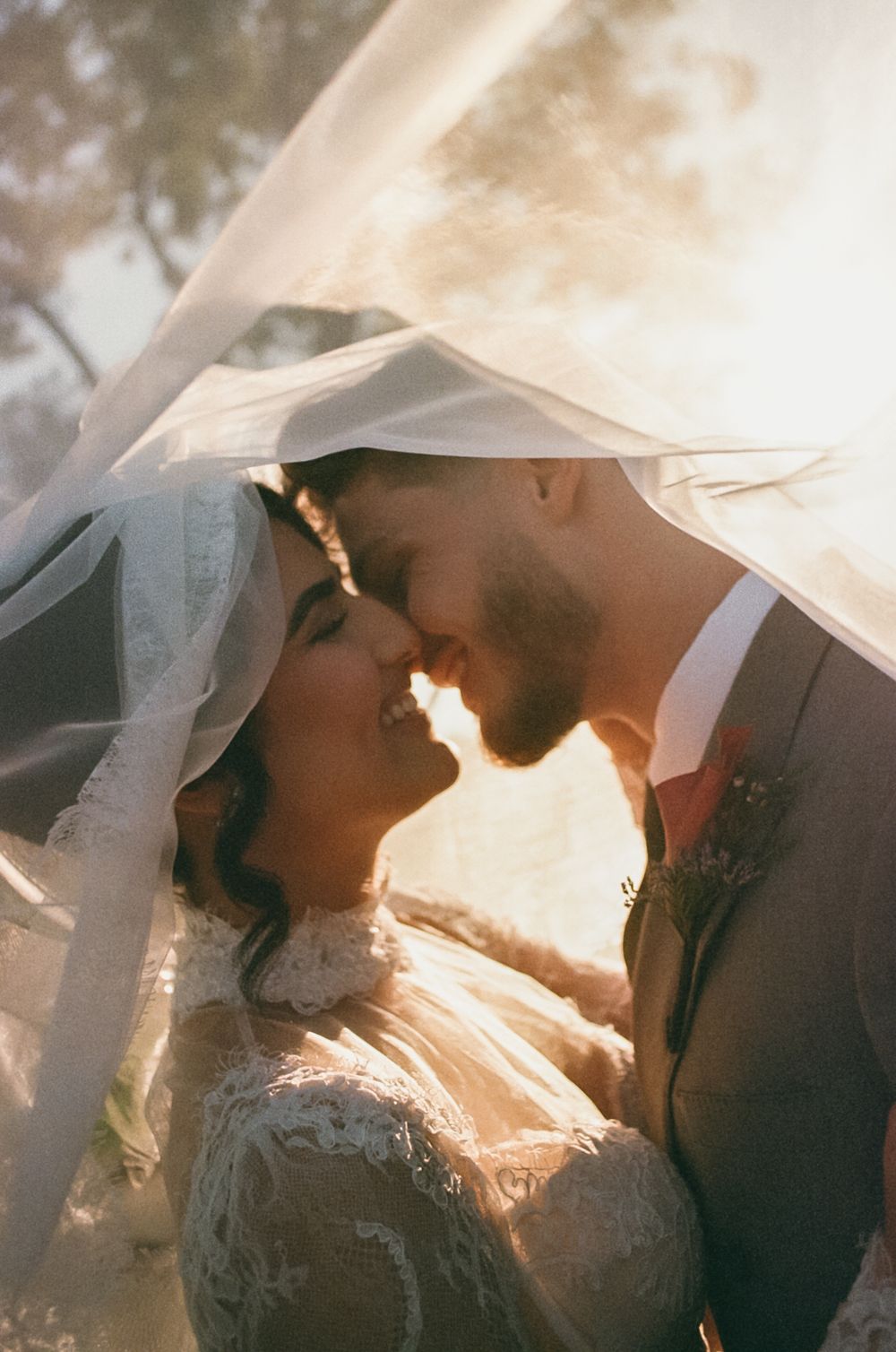 A romantic wedding moment captured under a veil with golden sunlight glowing through the delicate fabric.