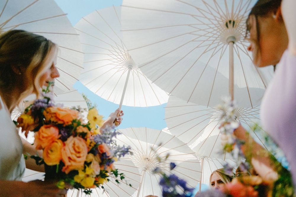 Wedding guests hold white parasols while gathered outdoors with colorful floral bouquets in a sunny garden setting.