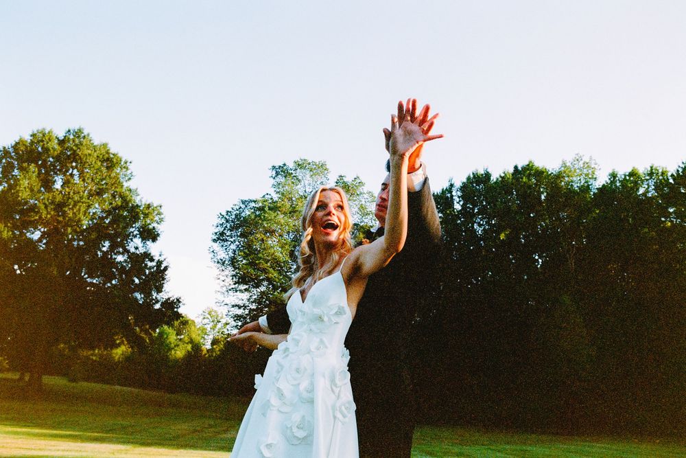Someone in a white dress jumps joyfully in a sunny park with trees in the background.