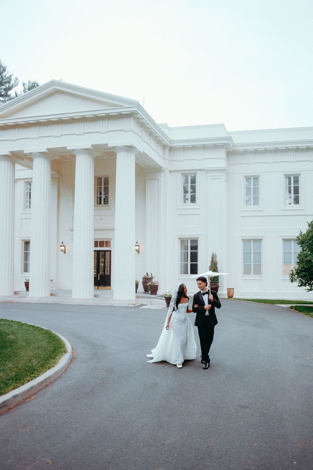 A couple walks along a curved driveway in front of an elegant white colonial-style building with columns.