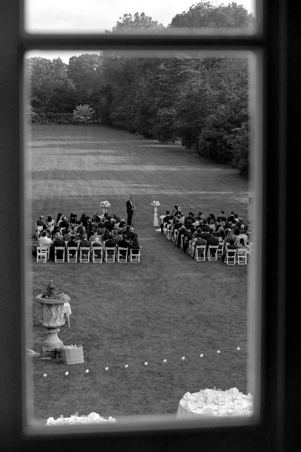 An outdoor wedding ceremony setup with white chairs arranged on a lawn viewed through a window.