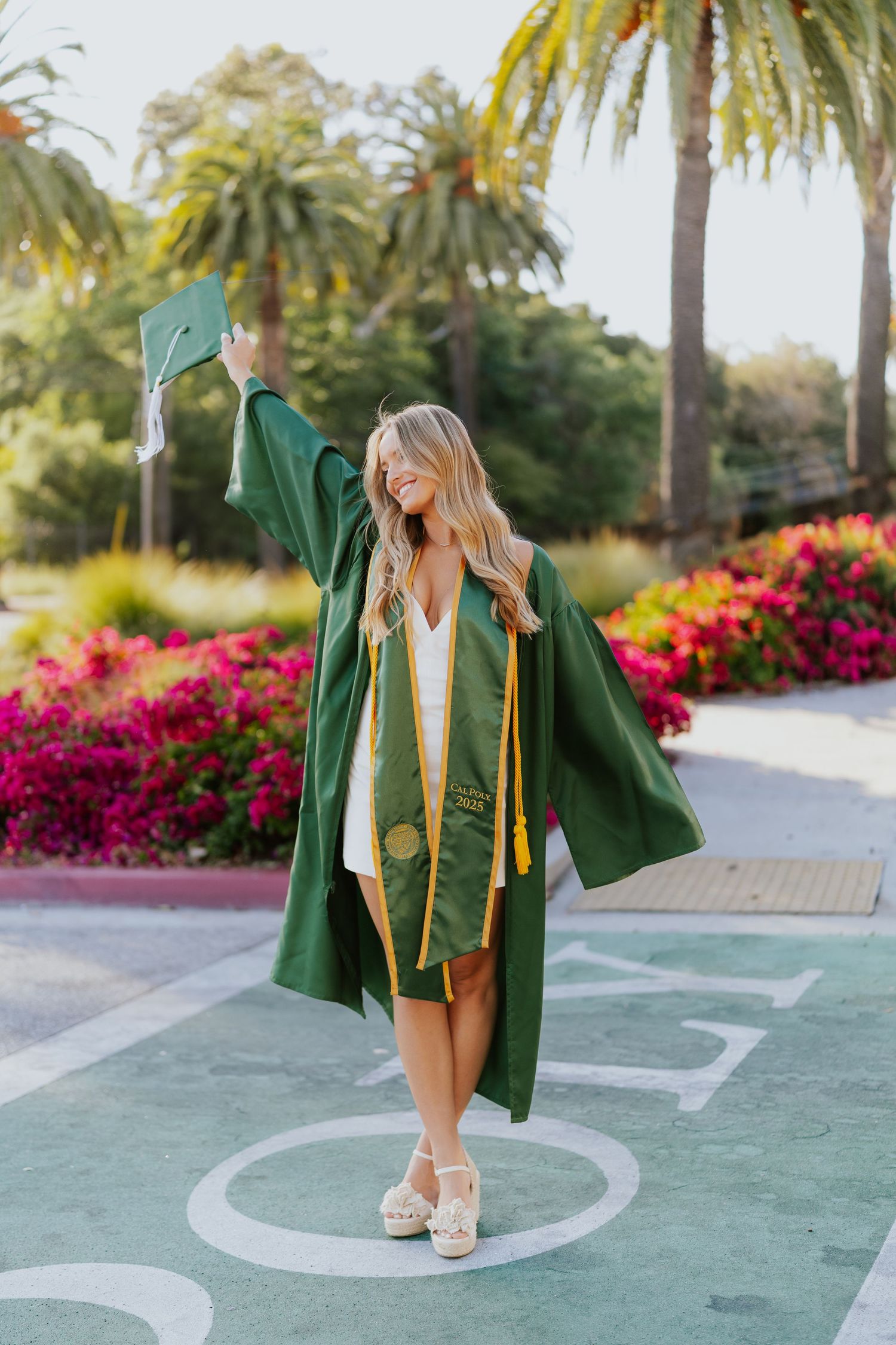 Graduate in green cap and gown celebrates on campus with palm trees and colorful flowers in background.