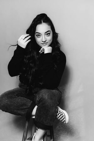 Black and white portrait of a person in dark sweater and checkered shoes sitting casually on a stool.