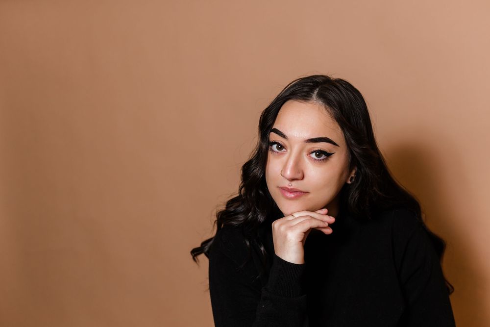 A person with dark hair and dramatic eye makeup poses against a brown background in a series of artistic portraits.