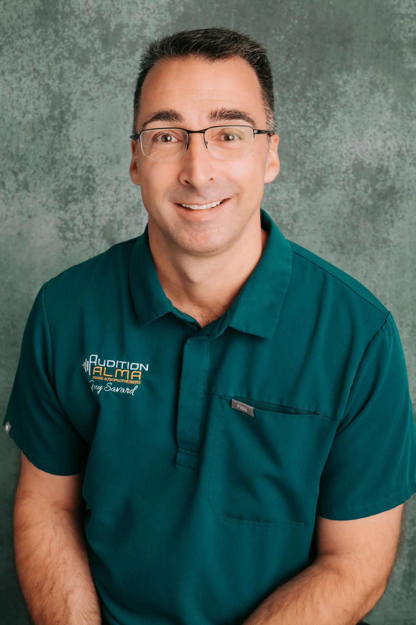 Professional headshot of someone wearing glasses and a green polo shirt with a company logo against a textured gray background.