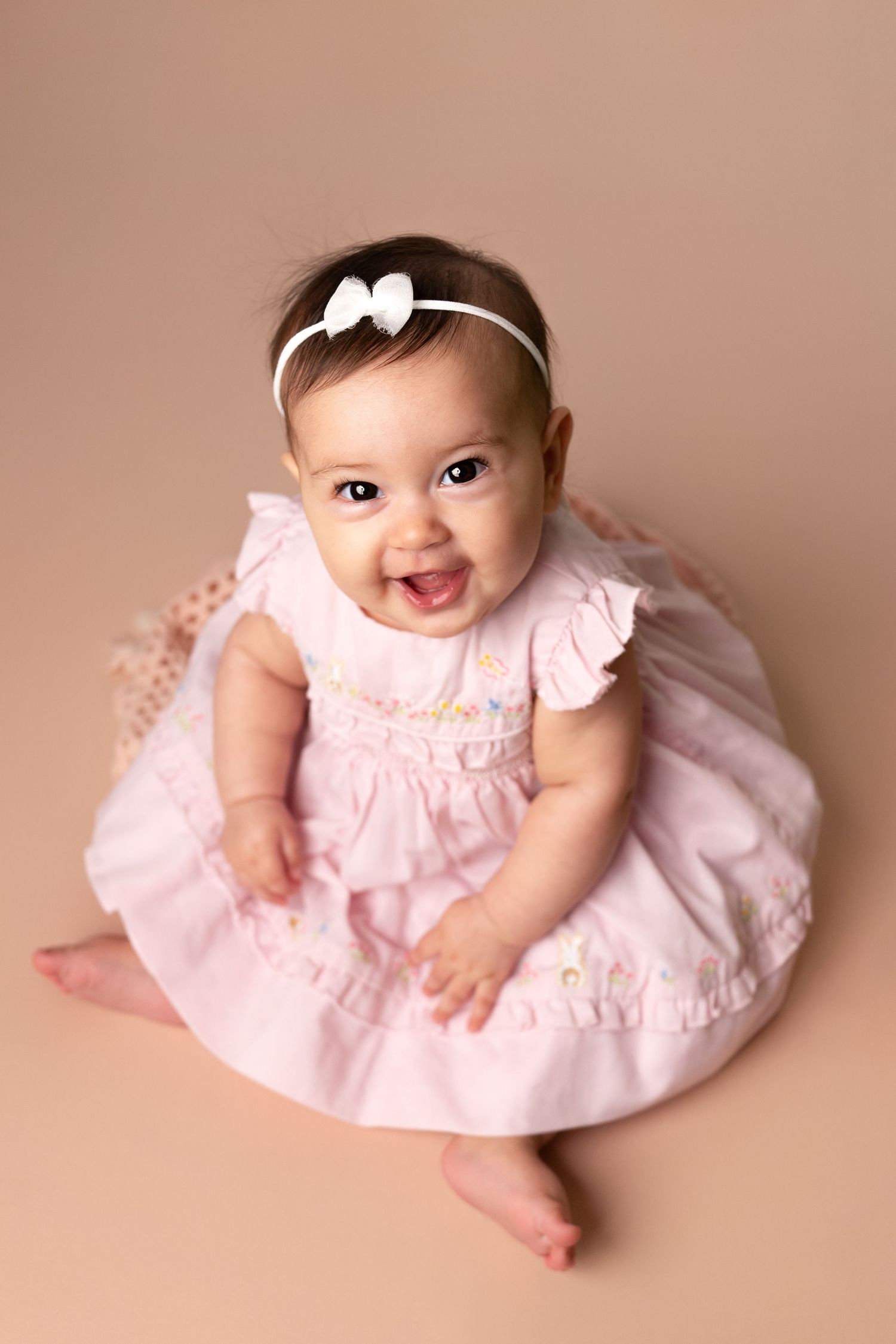 Baby wearing pink ruffled dress and white bow headband smiles joyfully while sitting on beige backdrop.