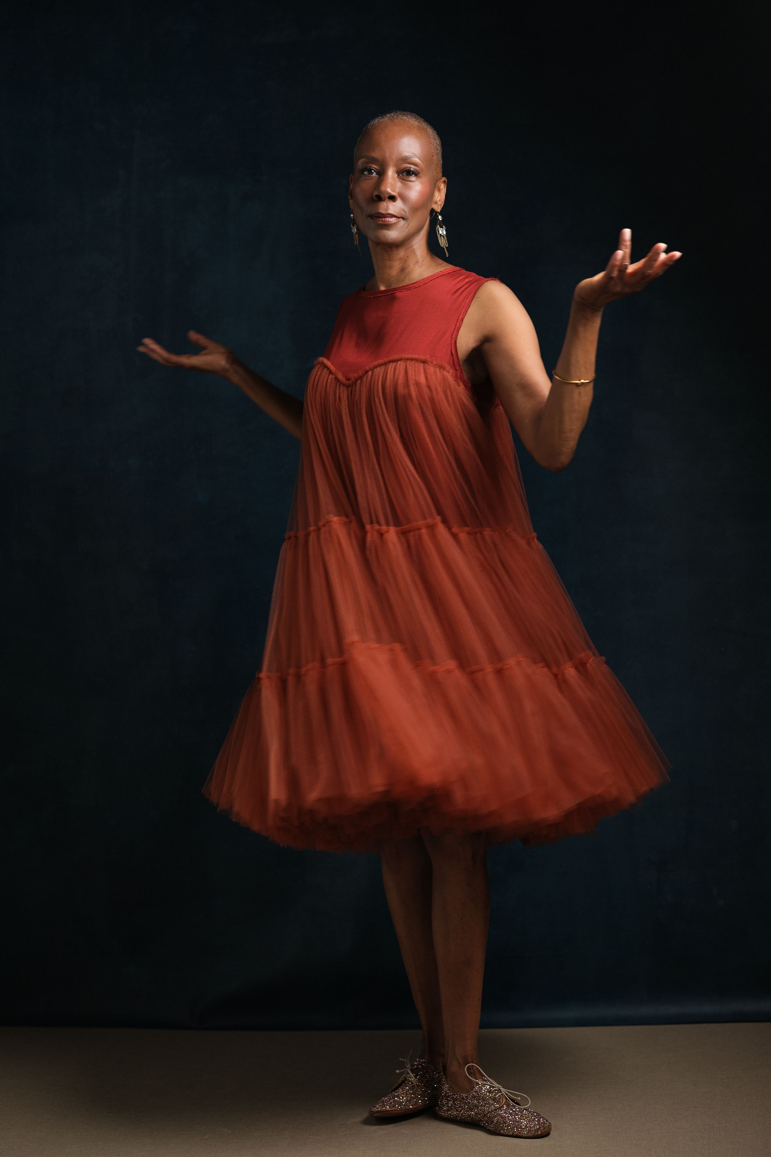 Graceful pose in red tiered dress with arms outstretched against dark wall.