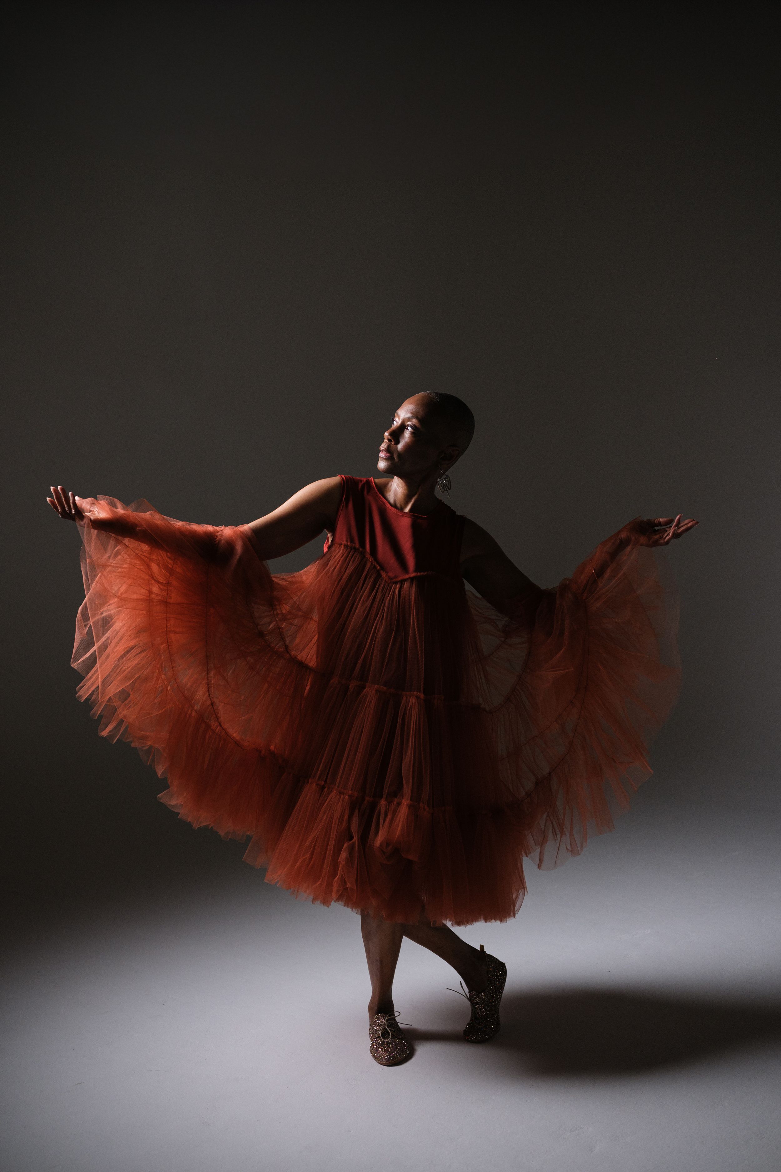 Dancer in flowing red tulle dress performs graceful movements in dramatic studio lighting.
