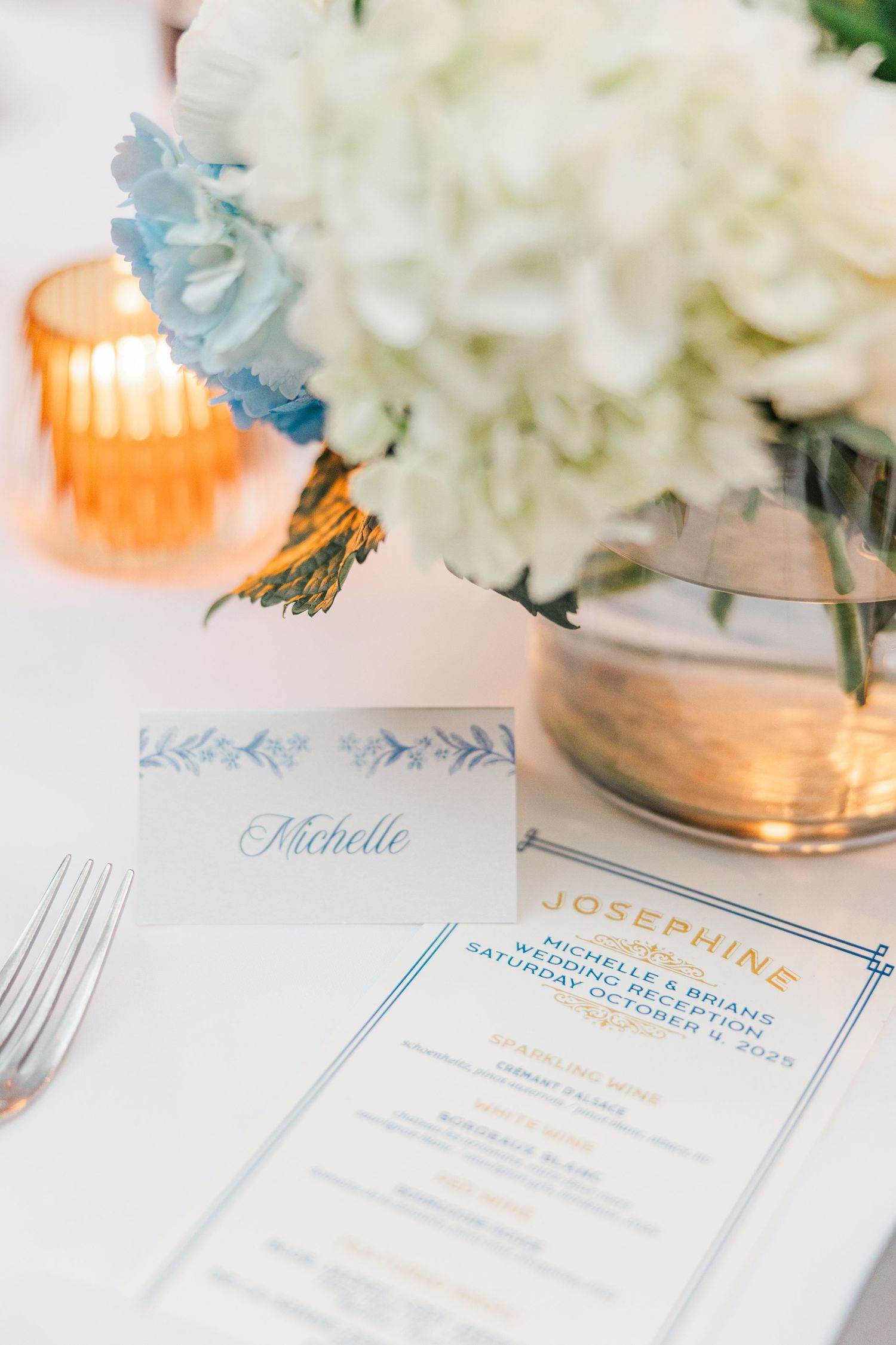 Elegant wedding invitation cards with blue calligraphy text displayed alongside white floral arrangements and soft lighting.