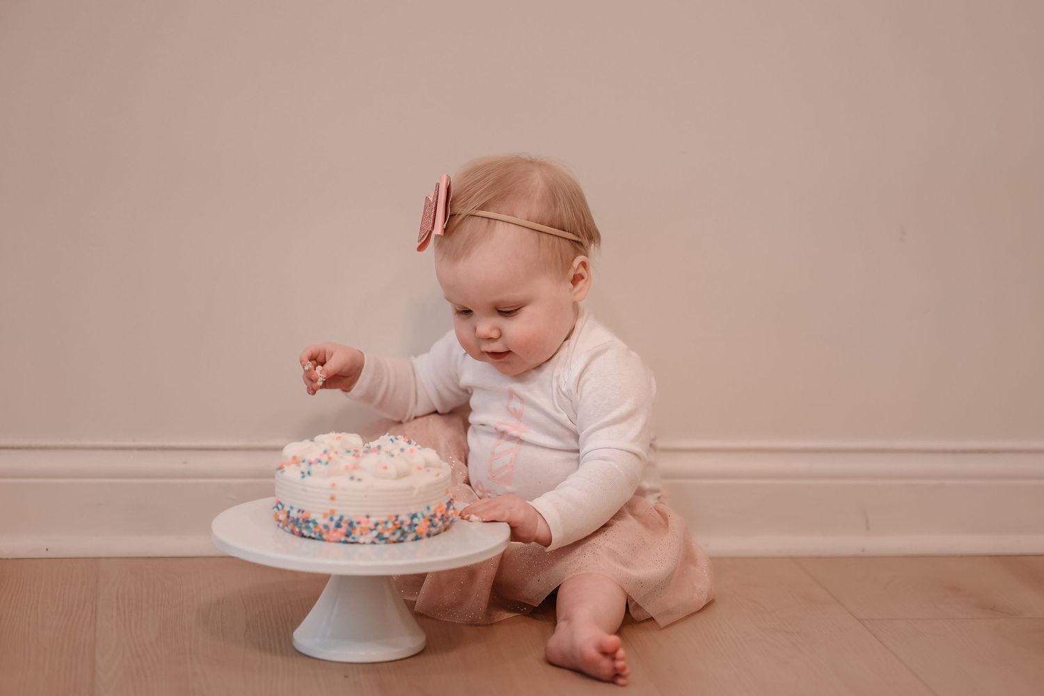 A small baby in white clothing reaches toward a cake on a white pedestal stand.