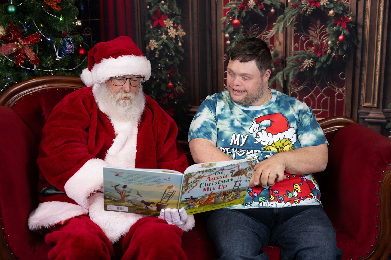 A festive holiday scene with someone in a red suit reading a book to a person in a blue shirt near a Christmas tree.