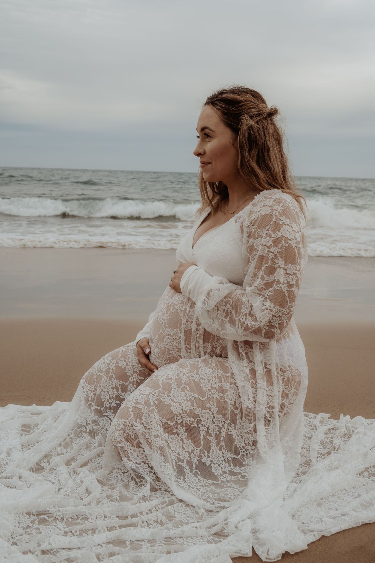Expectant mother in white lace dress poses gracefully on sandy beach during sunset at a central coast maternity photoshoot.
