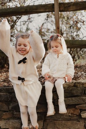 Two children in matching cream sweaters and tights sit together on rustic wooden steps during an outdoor photo session.