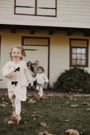 Children run and play on a grassy lawn in front of a white house with autumn leaves scattered on the ground.