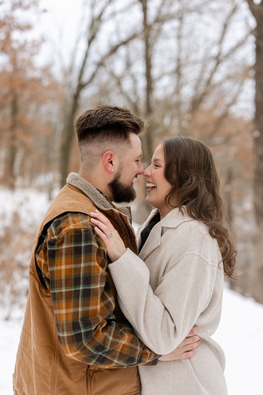 A romantic winter couple embraces in a snowy forest setting wearing plaid and cream colored outerwear.