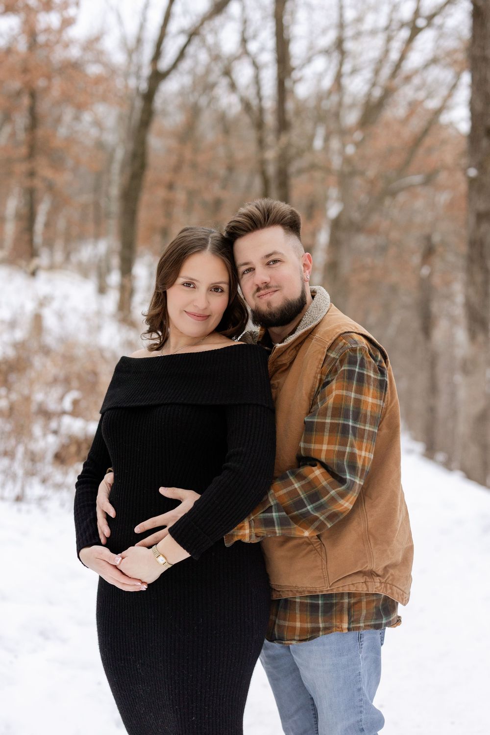 A couple poses together in a snowy winter forest setting wearing black and brown casual clothing.