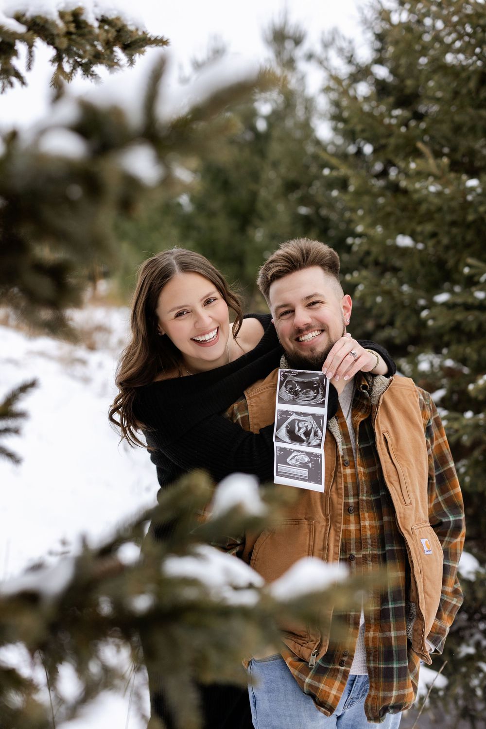 A couple stands among evergreen trees in winter while holding up an ultrasound photo together.