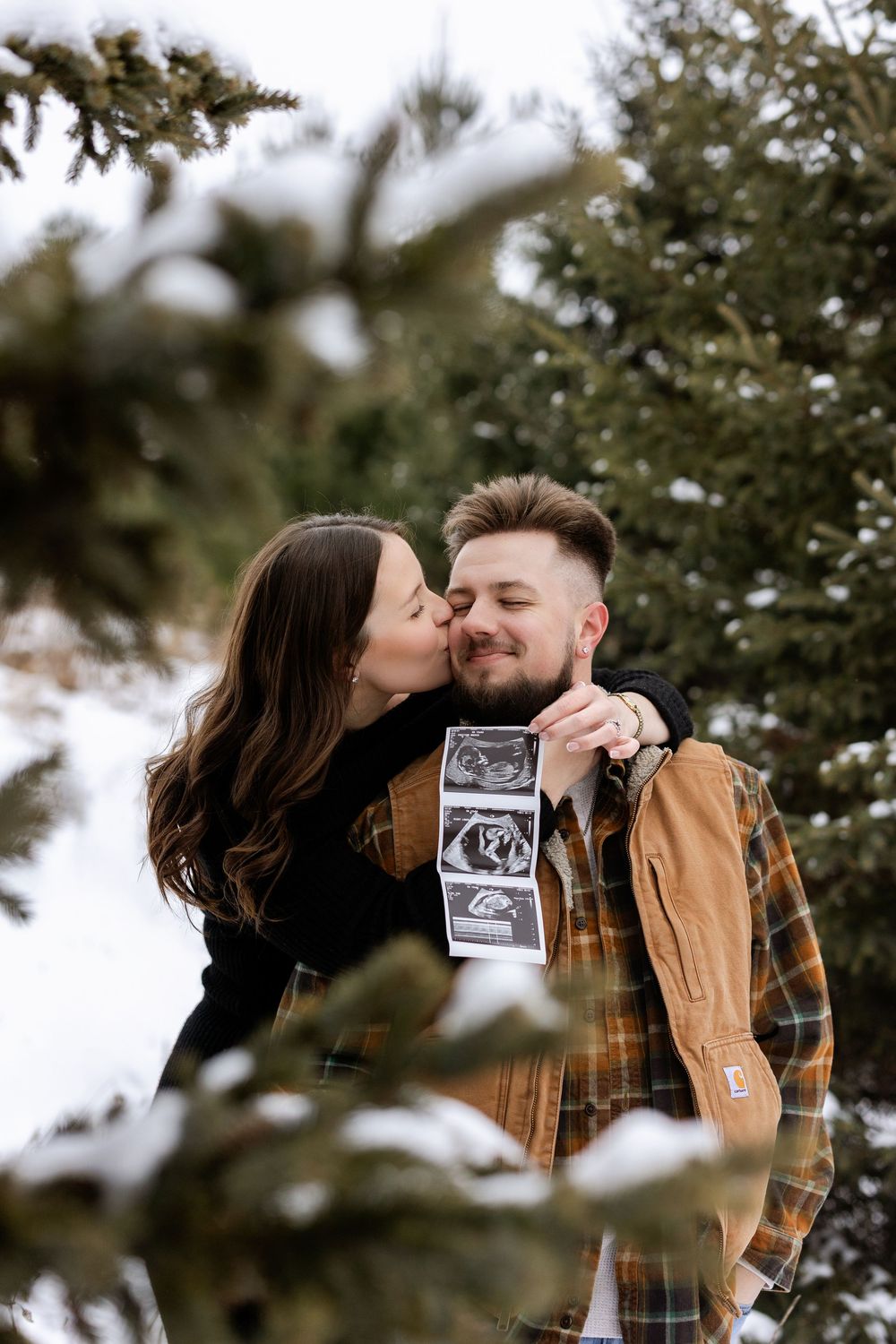 A couple shares a tender kiss while holding ultrasound photos in a snowy winter setting among evergreen trees.