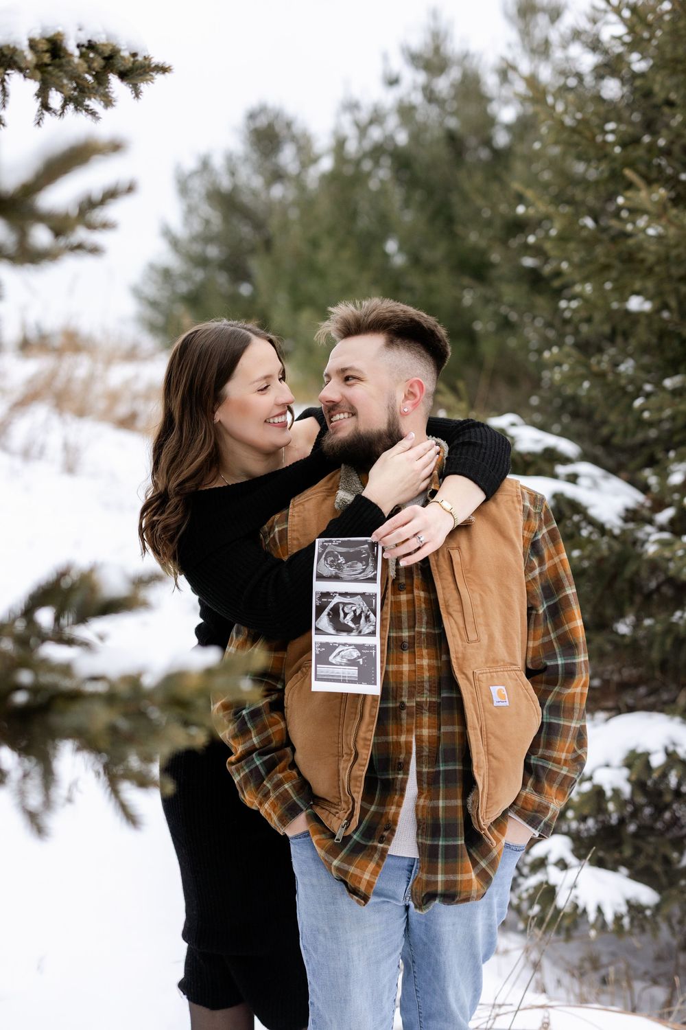 Couple sharing joyful moments in a snowy winter setting while holding ultrasound photos.