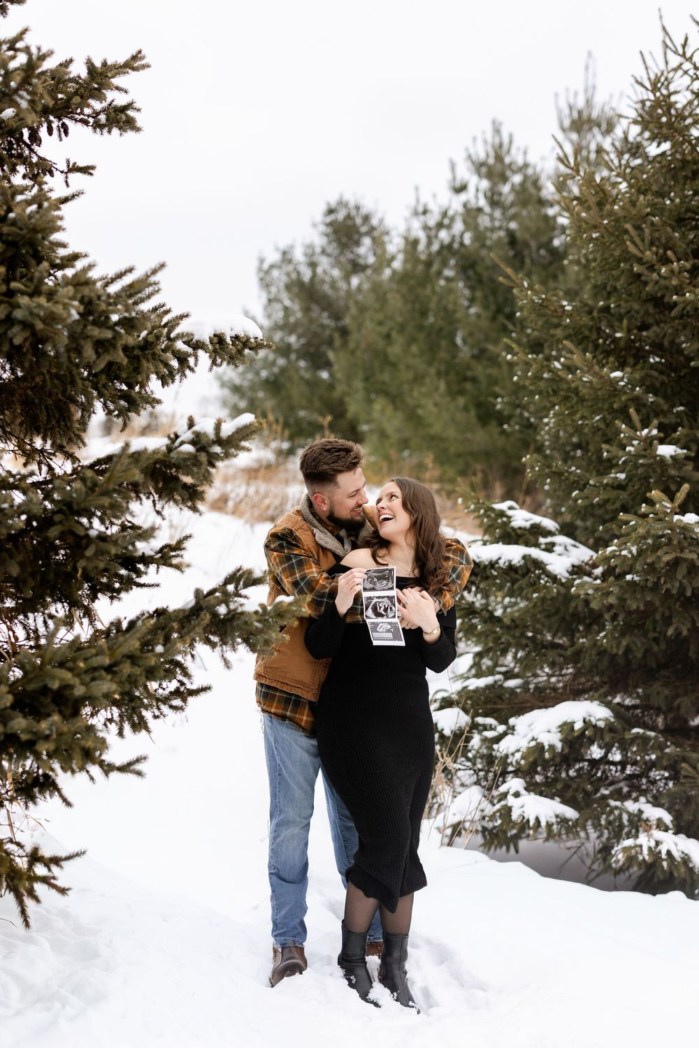 A couple shares romantic moments in a snowy winter setting with evergreen trees in the background.