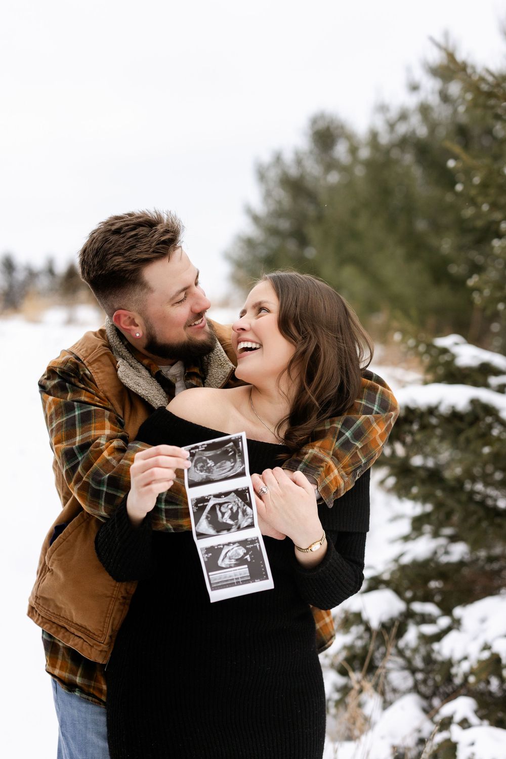 A couple embraces and laughs together in a snowy winter setting while holding a sonogram photo.