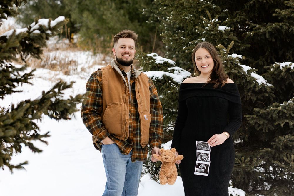 A couple poses together in a snowy winter setting with evergreen trees in the background.