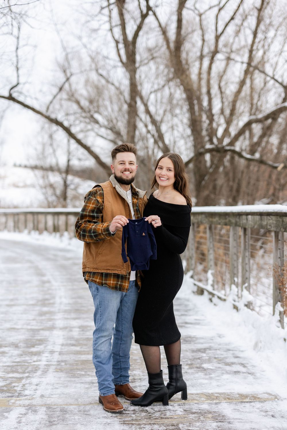 A couple poses together on a snowy path surrounded by bare winter trees.