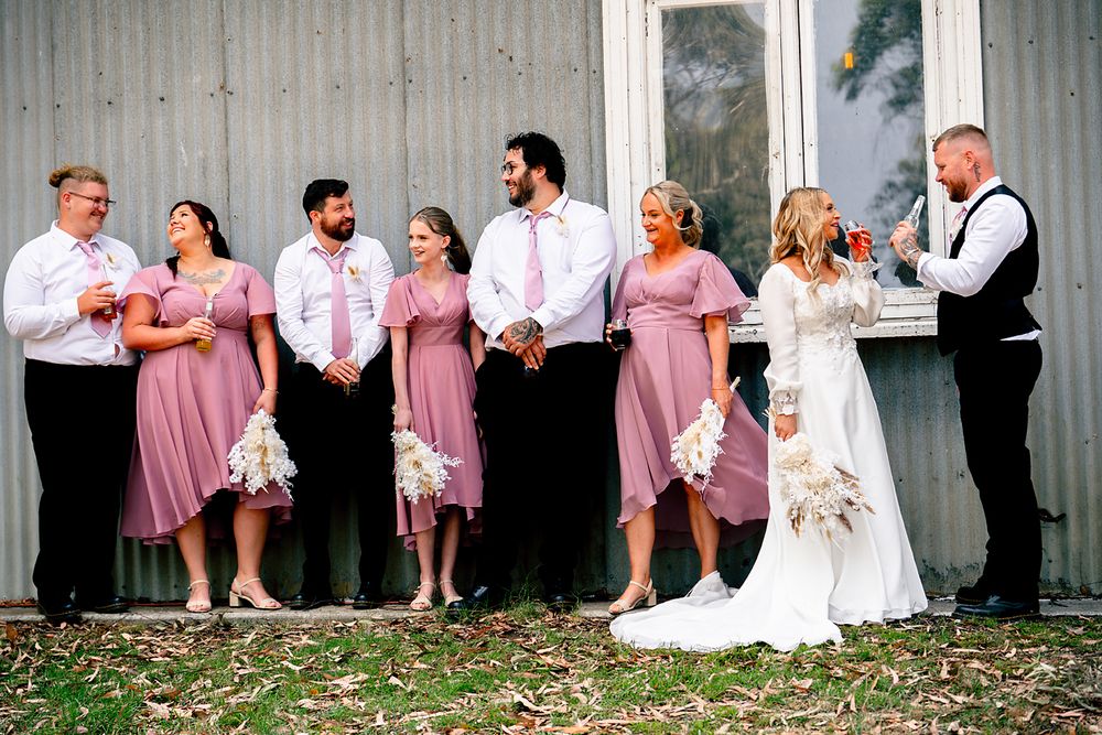 Wedding party poses together against a white barn wall wearing pink bridesmaid dresses and formal attire.