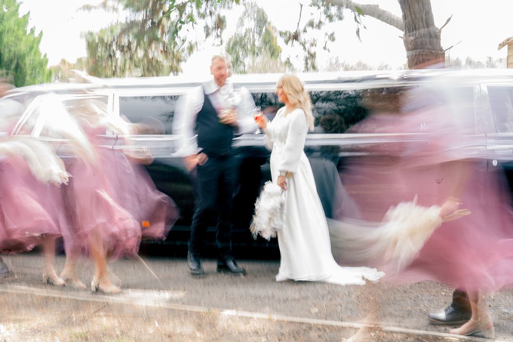 Wedding party members dance and twirl in a line showing off pink dresses and formal attire at an outdoor celebration.