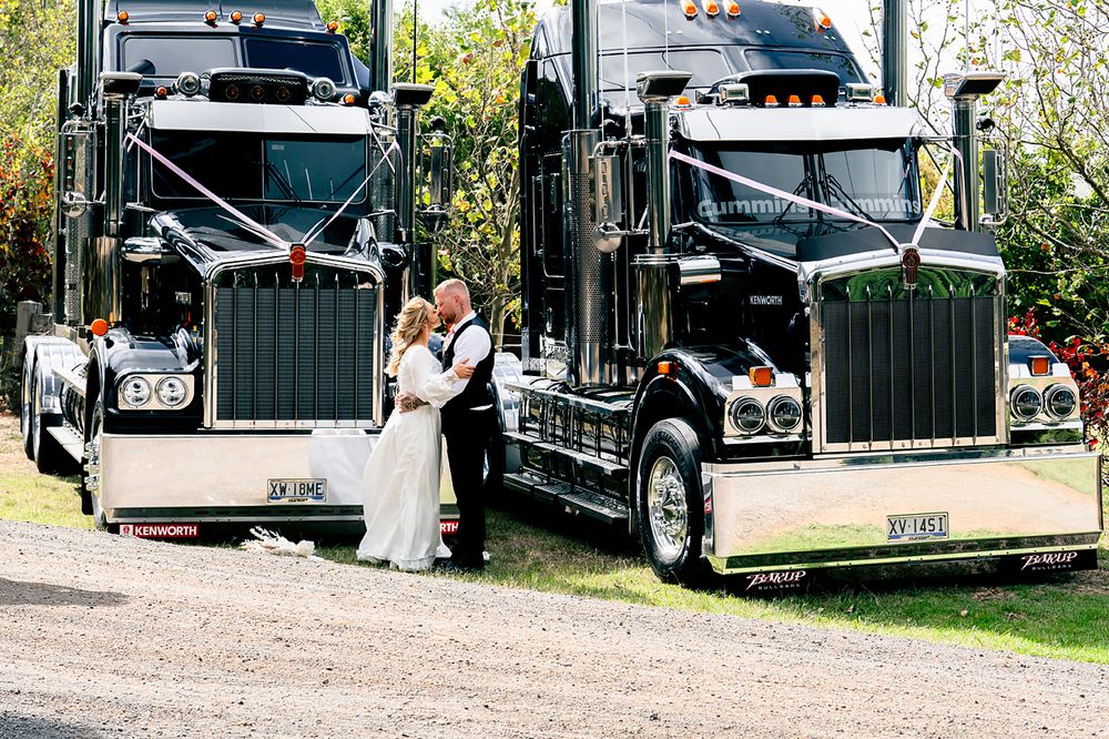 A series of black horse trailers parked in a line on a gravel path with trees and greenery in the background.