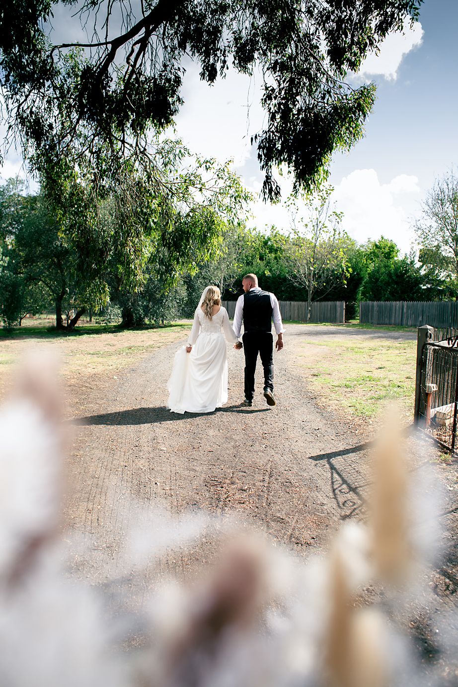 A couple walks together on a dirt path surrounded by trees and greenery on a sunny day.