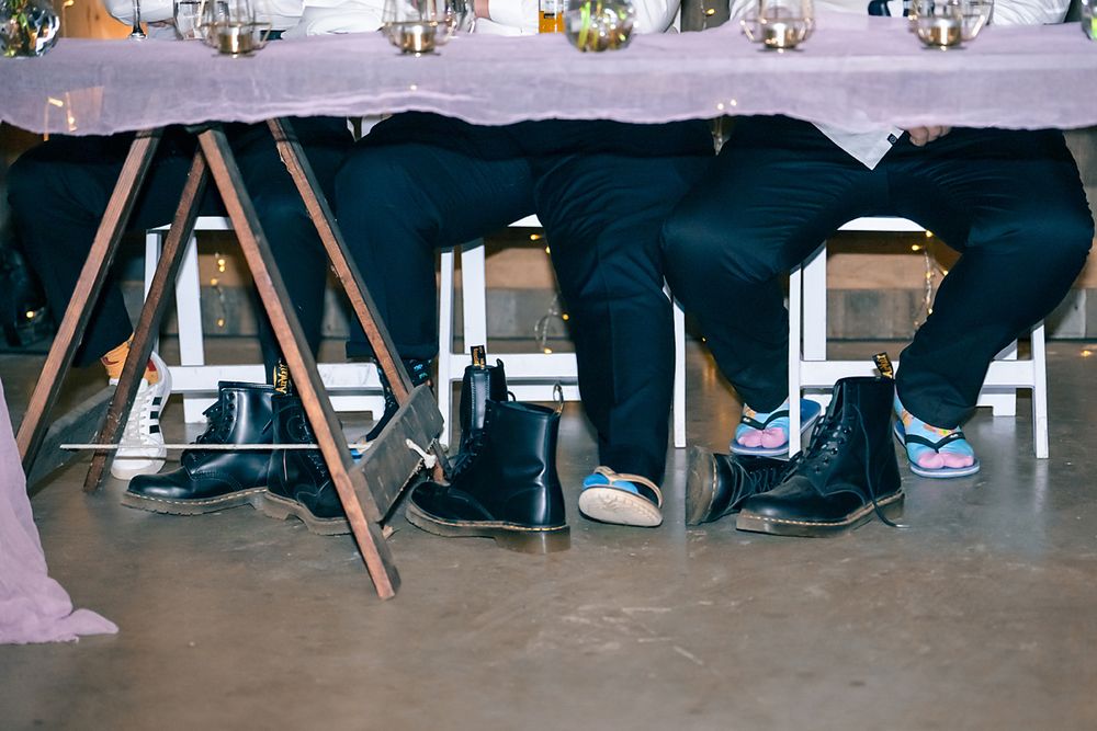 Wedding guests' feet and legs visible under a long banquet table draped with white linens at a reception.