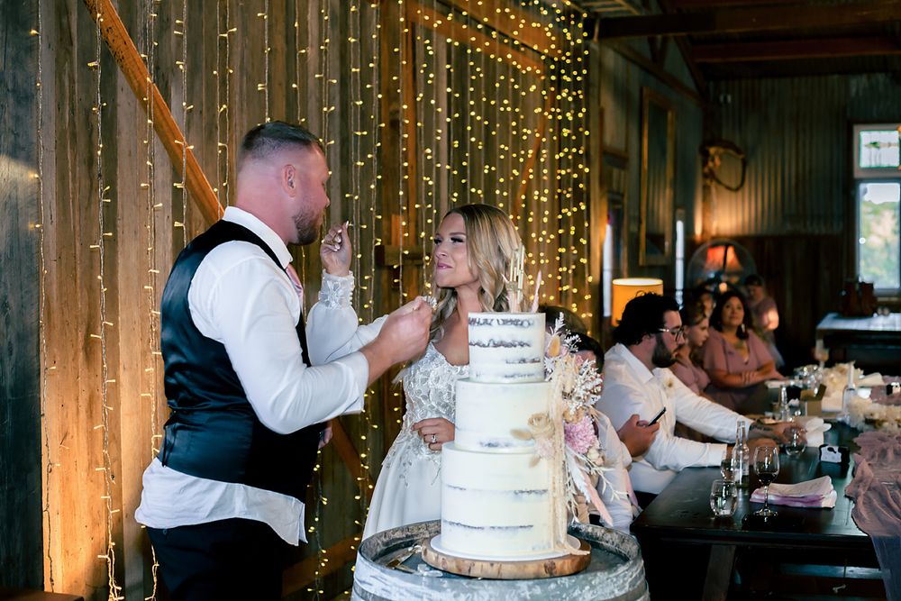 Wedding cake cutting ceremony in a rustic venue decorated with twinkling string lights.